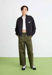 Giacca nera con zip e tasca sul petto, abbinata a pantaloni a gamba larga verde oliva e sneakers nere. Il modello si trova su un pavimento verde.