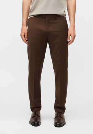 Mand iført mørkebrune slimfit habitbukser med frontfolder og brune lædersko mod hvid baggrund.