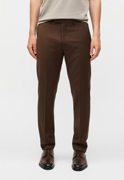 Homme portant un pantalon de costume slim fit marron foncé avec un pli sur le devant et des chaussures habillées en cuir marron sur fond blanc.