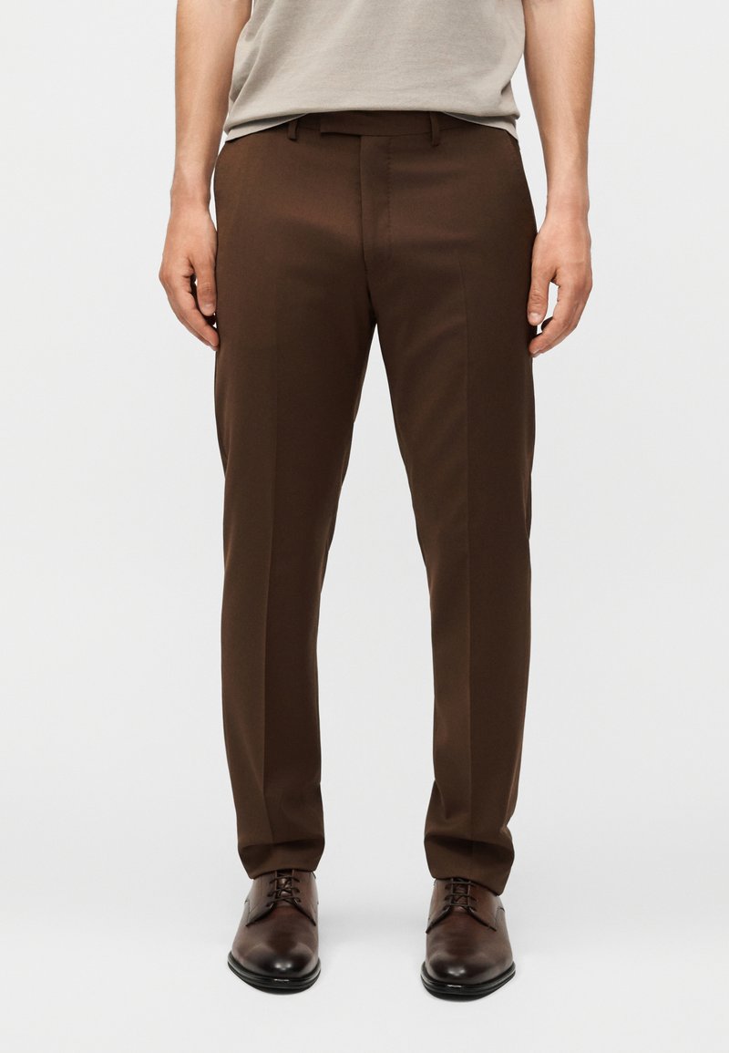 Άνδρας που φοράει σκουρό καφέ στενά παντελόνια με πιέτα μπροστά και καφέ δερμάτινα παπούτσια σε λευκό φόντο.