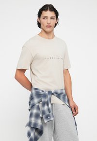 Lys beige t-shirt med et sort logo, parret med grå sweatpants og en blå ternet flannel skjorte bundet om taljen.