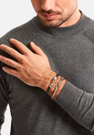 Drei Armbänder an einem Handgelenk: ein türkisfarbenes geflochtenes Lederband, eine silberne Kette und ein metallisches Stück mit rechteckigen Akzenten. Grauer Pullover im Hintergrund.