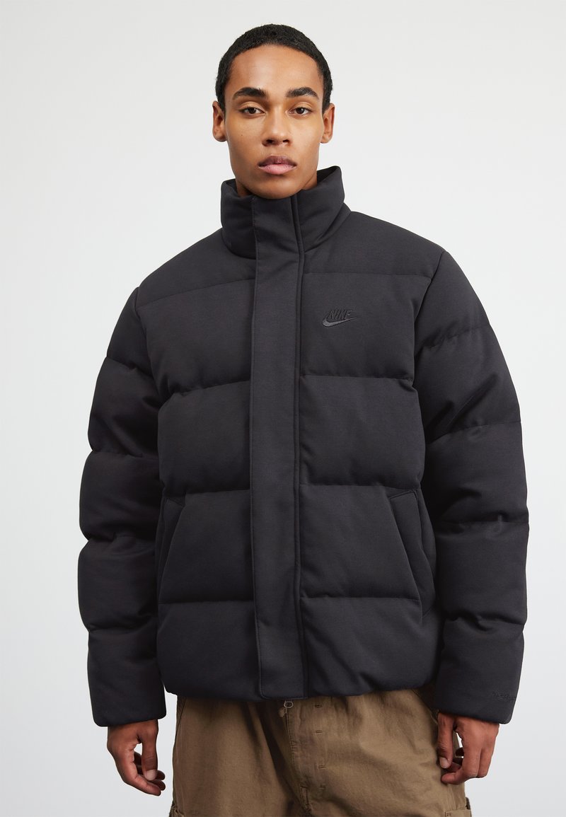 Geacă puffer neagră, fabricată dintr-un material moale și căptușit, cu un guler înalt, având un logo Nike discret pe piept și buzunare laterale.