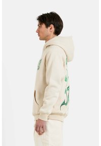Felpa beige con tasca frontale, caratterizzata da grafiche verdi sui lati. Il materiale sembra morbido con un fit rilassato e un cappuccio regolabile con cordino.