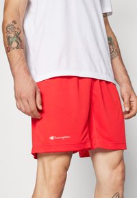 Κόκκινες δικτυωτές αθλητικές σορτς με υφή, που διαθέτουν το λογότυπο "Champion" σε λευκό στην κάτω ήμισυ και ελαστική μέση.