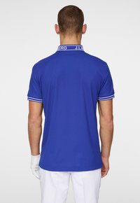 Blue polo shirt with a tailored fit. Features a ribbed collar with white detailing and a branding label at the back. Short sleeves.