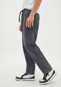 Černé džíny z denimu s rovnými nohavicemi, vybavené stahovací šňůrkou v pase a bočními kapsami, v kombinaci s černobílými kotníkovými teniskami.