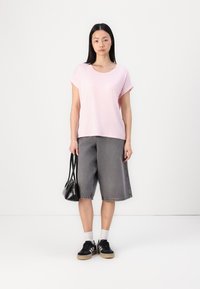 Lyserosa stripete t-skjorte med kort erm, løse grå denimshorts, svarte joggesko med beige såler, holder en liten svart håndveske.