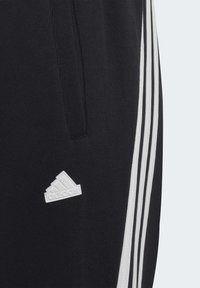 Pantalones deportivos negros con tres franjas verticales blancas y un pequeño logo de Adidas en blanco en el lado izquierdo. Textura suave, diseño casual.