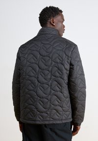 Kvilta melna jaka ar paaugstinātu rakstu, standarta apkakli un elastīgiem aprocēm. Tekstūra izskatās gluda ar vieglu dizainu.