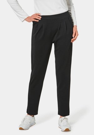 Schwarze plissierte Hose mit breitem Bund, schmal zulaufendem Bein und glatter Stofftextur. Getragen mit weißen Sneakers für einen lässigen Look.