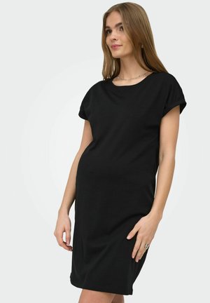 Femme portant une robe noire simple, à manches courtes et longueur genou, debout devant un fond uni gris clair, avec peu de bijoux.