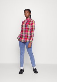 Camisa quadriculada em vermelho e verde, com botões, duas bolsos no peito, combinada com jeans azul claro e mocassins pretos, sobre um fundo simples.