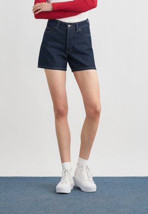 Žena oblečená v tmavomodrých riflových šortkách, bielych vysokých teniskách a červenom dlhom rukáve, so založenými rukami.