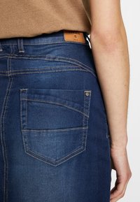 Bakifrån vy av person som bär mörkblå denimjeans med detaljerad sömnad och brun lädermärke, i kombination med en ljusbrun topp.