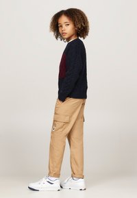 Maglione blu navy a treccia con dettagli bordeaux, pantaloni cargo beige con tasche laterali, sneakers bianche con dettagli blu e rossi, e capelli texturizzati.