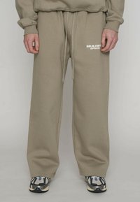 Beige Jogginghose mit einem Tunnelzug in der Taille, gerippten Bündchen und einem weißen Logo am linken Oberschenkel. Das Material wirkt weich und strukturiert.