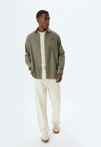 Olivgrünes Button-Up-Hemd aus leichtem Stoff, mit Vorderasche, kombiniert mit cremefarbenen weiten Hosen und beigefarbenen Sneakers. Einfaches, lässiges Design.