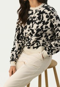 Femme assise sur un tabouret en bois, portant un pantalon beige et un pull à manches longues à motif abstrait noir et blanc.