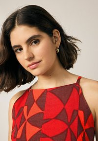 Rot und orange gemustertes Kleid mit Halterneck und dünnen Trägern, aus leichtem Stoff gefertigt, mit geometrischen Formen und kontrastierenden Farben.