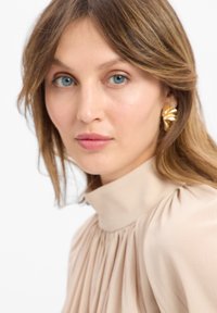 Blusa beige con collo alto e dettagli plissettati. Orecchini d'oro dichiarativi aggiungono contrasto. Texture morbida con trucco sobrio e dall'aspetto naturale.