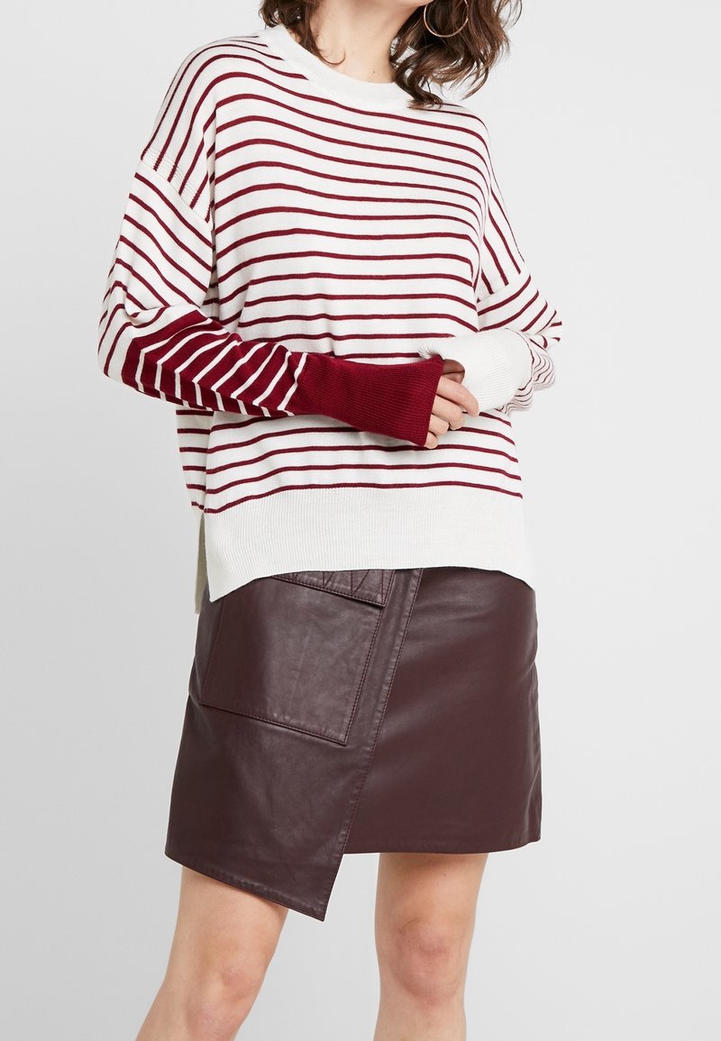 Femme portant un pull rayé blanc et bordeaux à manches longues et une jupe en cuir asymétrique marron foncé avec une poche avant.