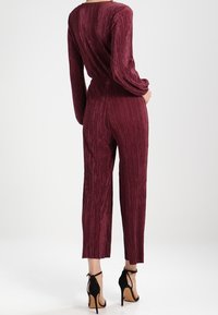 Burgundy jumpsuit with vertical ribbed texture, long sleeves, and tapered pants, paired with black strappy high-heeled sandals.