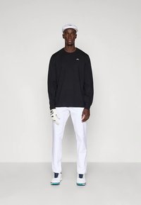 Ψηλός άνδρας που φοράει μαύρο μακρύ μανίκι, λευκά παντελόνια, λευκό γάντι γκολφ στο αριστερό χέρι, λευκό καπέλο και λευκά παπούτσια γκολφ με μπλε κορδόνια.