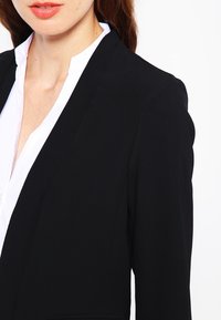 Mustat bleiseri, jossa on muotoiltu leikkaus, sileä kangas, terävät läpät ja yksivärinen malli, yhdistettynä valkoiseen kauluspaitaan.