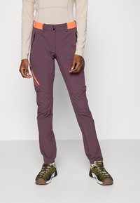 Pantalones exteriores morados con una cintura naranja, bolsillos con cremallera y costuras en panel; combinados con zapatos deportivos marrones y verdes.