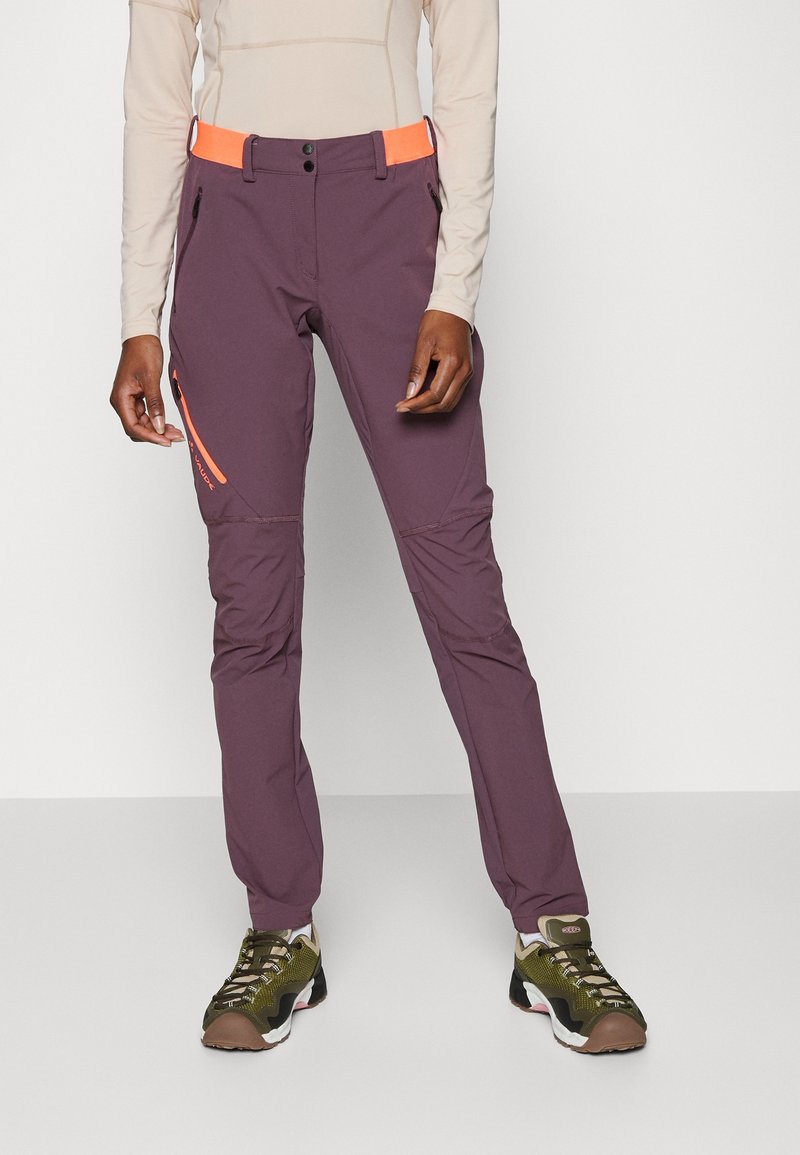 Pantalones exteriores morados con una cintura naranja, bolsillos con cremallera y costuras en panel; combinados con zapatos deportivos marrones y verdes.