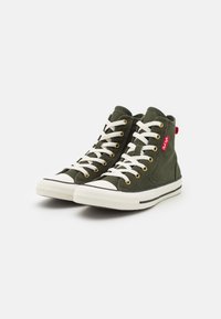 Un paio di sneakers alte in tela verde oliva con suole e lacci in gomma bianca, occhielli dorati e un'etichetta in tessuto rossa su sfondo bianco.