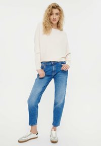 Lichtroze geribbelde trui, los model. Blauwe spijkerbroek met een rechte snit. Witte sneakers met grijze accenten en een bruine zool.