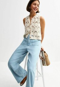 Chaleco de ganchillo en blanco roto con patrones florales y botones, combinado con jeans de pierna ancha en azul claro y sandalias marrones. Bolsa tejida con logo.