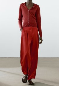 Osoba ubrana w czerwony kardigan z guzikami, luźne czerwone spodnie z zakładkami i czarne sandały, idąca po gładkiej podłodze wzdłuż gładkiej białej ściany.