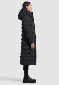 Longo cappotto imbottito nero con cappuccio, caratterizzato da un design trapuntato, chiusura zip e due tasche laterali. Abbinato a stivali neri alla caviglia.