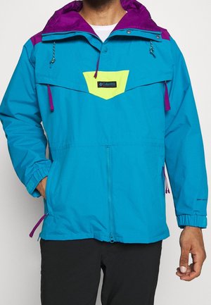Mann trägt türkis-violette Kapuzenjacke mit gelbem Brustaufnäher und Columbia-Logo, Hände in den Taschen vor weißem Hintergrund.