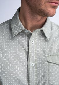 Camisa de botón verde claro con una tela texturizada, que presenta un pequeño patrón geométrico y un bolsillo frontal. Botones y detalles del cuello en blanco son visibles.