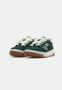 Grüne Wildleder-Sneaker mit weißen Schnürsenkeln, ausgestattet mit einer Gummisohle und einer strukturierten braunen Außensohle. Auffälliges Logo an der Seite.