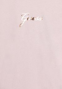 T-shirt rosa chiaro realizzato in morbido cotone, caratterizzato da un grande logo dorato "Guess" in un font corsivo sul petto.