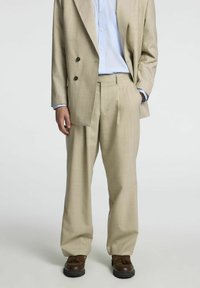 Bež žakard blazer s črnimi gumbi, v kombinaciji z usklajenimi nabranimi hlačami in svetlo modro prugasto srajco. Rjave čevlje.