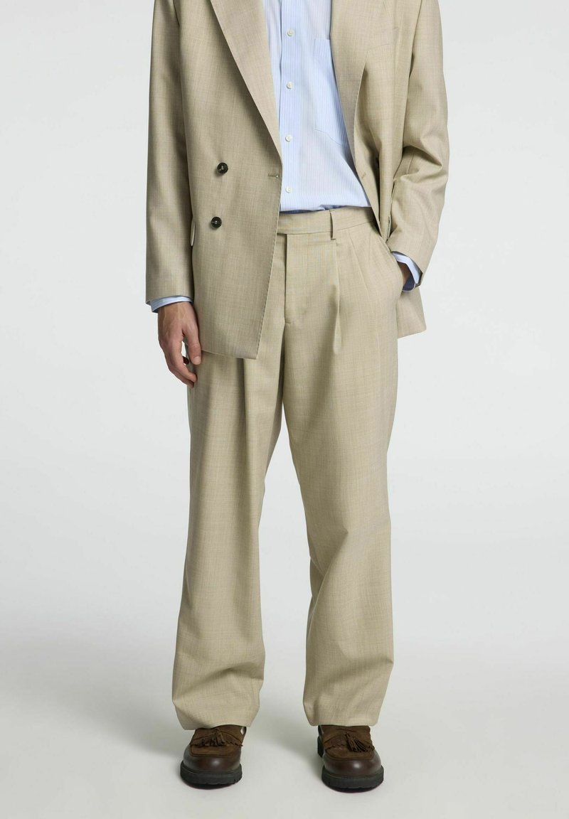 Bež žakard blazer s črnimi gumbi, v kombinaciji z usklajenimi nabranimi hlačami in svetlo modro prugasto srajco. Rjave čevlje.