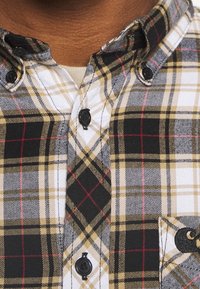 Camicia a quadri in cotone, caratterizzata da un mix di motivi neri, bianchi, gialli e rossi. Notabile per il colletto e il dettaglio della tasca a contrasto.