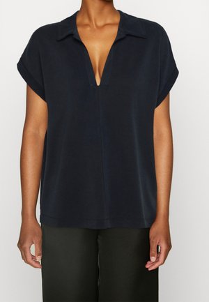 Femme portant un chemisier ample bleu marine à manches courtes avec un décolleté en V profond et un col, assorti à un pantalon foncé.