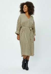 Lange, beige-zwart geblokte jurk met een ceintuur rond de taille, V-halslijn en brede mouwen, gecombineerd met zwarte enkellaarsjes.