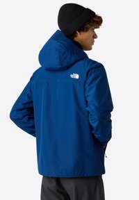 Mladý člověk v modré bundě North Face s kapucí a černou čepicí, stojící v profilu s rukama v kapsách.