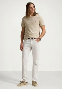 Męski model z długimi włosami, ubrany w beżową koszulkę, białe dżinsy, beżowe trampki i zegarek na nadgarstku, stojący z jedną ręką w kieszeni.