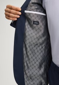 Giacca da abito blu navy, con foderatura a motivi grigi e etichetta "STRETCH TECHNOWOOL". I bordi bianchi accentuano l'interno.