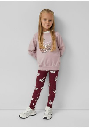Jong meisje staat tegen een grijze muur en draagt een roze sweatshirt met een gezwinde zwaan en bordeauxrode leggings met witte zwaanprints.