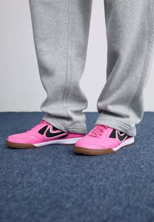 Person iført grå joggingbukser og lyserøde sportssko står på blå tæppebelagt gulv.